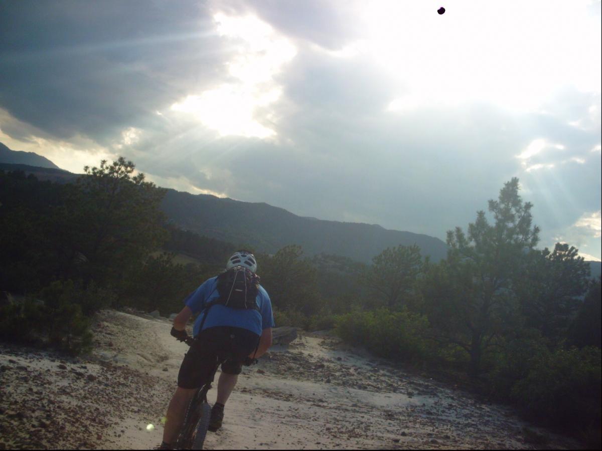 A mountain biker navigating a rocky trail surrounded by trees, with sunlight breaking through cloudy skies in the background. Ute Valley Park mountain bike trail.