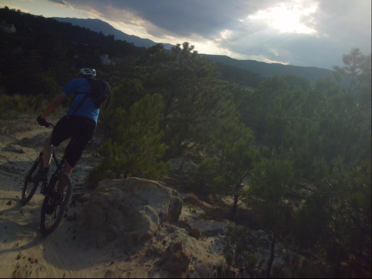 A mountain biker riding along a rocky trail, surrounded by lush greenery and distant mountains under a cloudy sky. Ute Valley Park mountain bike trail.