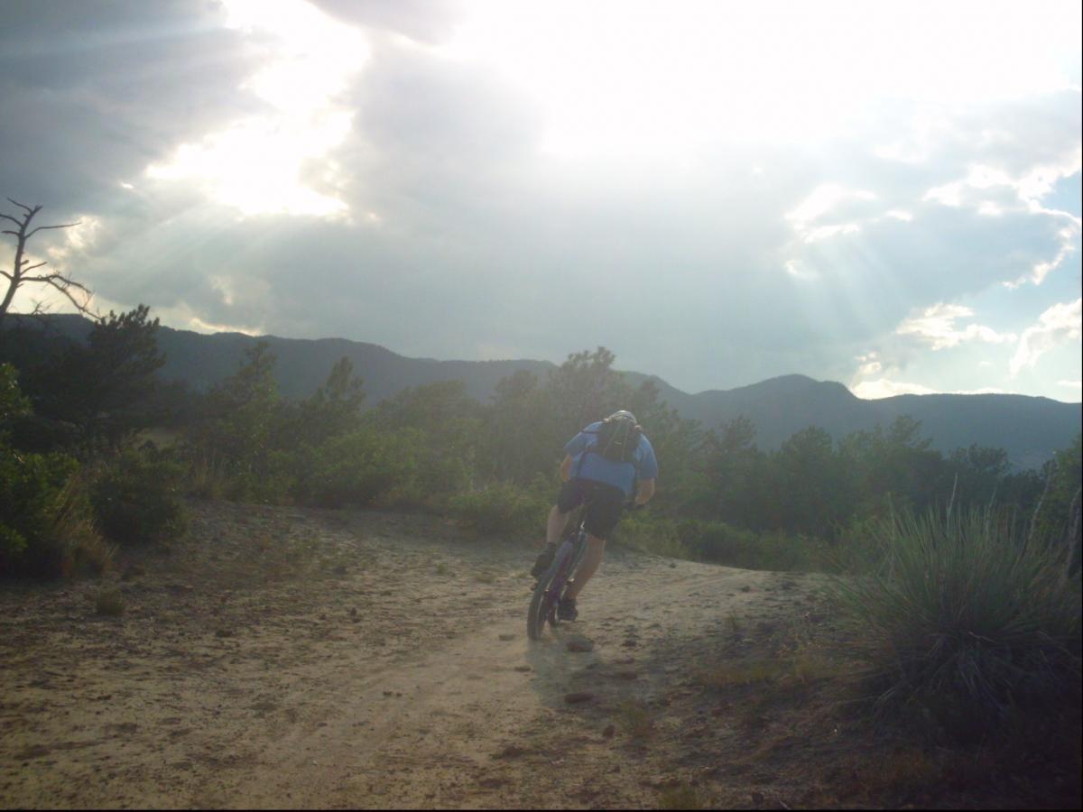 A cyclist riding on a dirt trail surrounded by trees and mountains, with sunlight breaking through the clouds in the background. The setting suggests a scenic outdoor adventure, showcasing the beauty of nature. Ute Valley Park mountain bike trail.