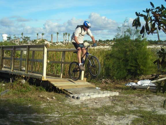A cyclist wearing a helmet and protective gear is performing a jump off a wooden bridge located in a natural outdoor setting with tall grass and trees in the background. Quiet Waters Park mountain bike trail.