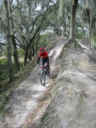 A cyclist wearing a red shirt and helmet rides a mountain bike along a dirt trail bordered by trees with Spanish moss. The path is narrow and elevated, indicating a rugged terrain in a forested area. Loyce E. Harpe Park mountain bike trail.