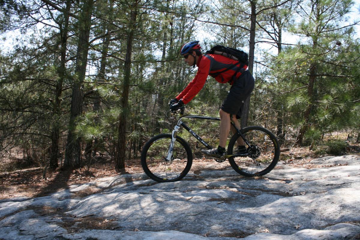 A cyclist riding a mountain bike over rocky terrain in a wooded area, wearing a red long-sleeve shirt, black shorts, a helmet, and gloves. The background features trees and sunlight filtering through the foliage. Georgia International Horse Park mountain bike trail.
