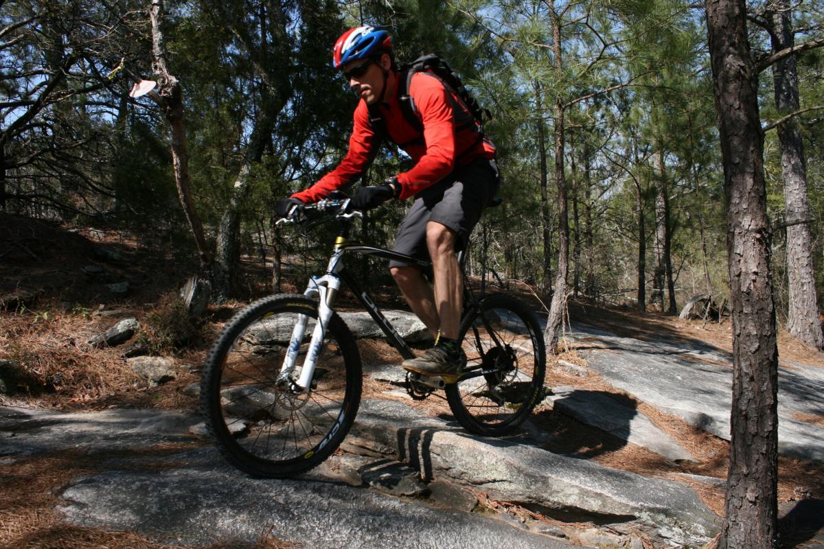 A person riding a mountain bike over rocky terrain in a forested area. The cyclist is wearing a red long-sleeve shirt, black shorts, a helmet, and gloves, with a backpack on their back. Sunlight filters through the trees, highlighting the natural setting. Georgia International Horse Park mountain bike trail.