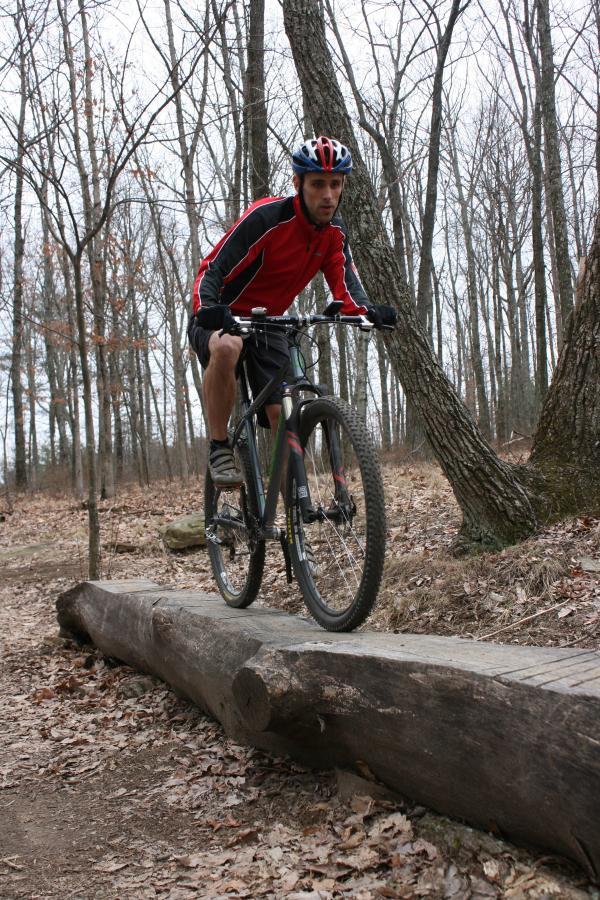 A mountain biker in a red and black jersey rides a bicycle along a wooden log placed on a trail in a wooded area, surrounded by bare trees and fallen leaves. The rider is focused and balanced as he navigates the obstacle. Raccoon Mountain Trail Network mountain bike trail.
