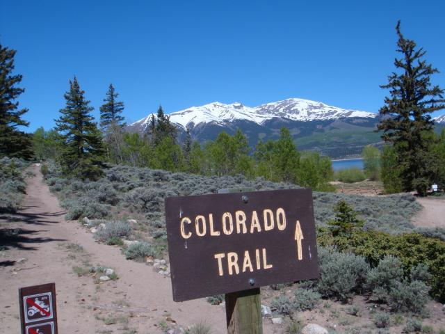 A wooden sign indicating "Colorado Trail" with an arrow pointing upward, surrounded by a scenic landscape of mountains, trees, and a clear blue sky. The path leading from the sign is lined with shrubs and rocks, suggesting a hiking trail in a natural outdoor setting. Snow-capped mountains can be seen in the background. Colorado Trail mountain bike trail.