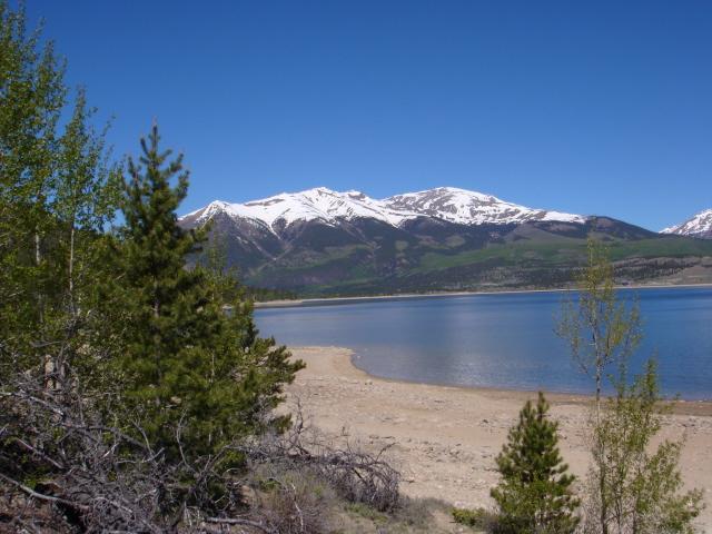 A scenic view of a lake surrounded by mountains, with snow-capped peaks in the background. The foreground features a sandy shoreline and patches of greenery, including coniferous trees. The sky is clear and blue, creating a tranquil atmosphere. Colorado Trail mountain bike trail.