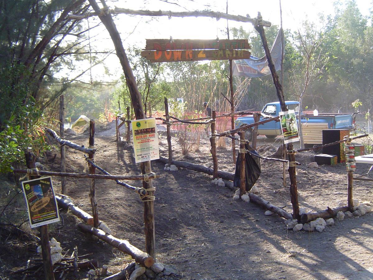 Entrance to a rustic pathway marked by a wooden sign that says "Down 2 Earth." The path is framed by rustic wooden posts and ropes, with information posters attached to the left side. In the background, a blue pickup truck and various structures are visible amidst sparse vegetation and dirt. The scene is illuminated by natural sunlight. Oleta River State Park mountain bike trail.