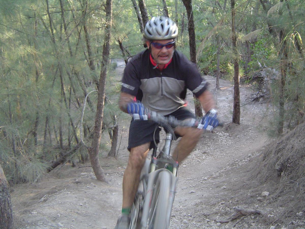 A mountain biker navigating a dirt trail surrounded by trees, with a focus on the rider's expressive face and dynamic riding posture. The cyclist wears a helmet and cycling gear, showcasing an adventurous outdoor activity. Oleta River State Park mountain bike trail.
