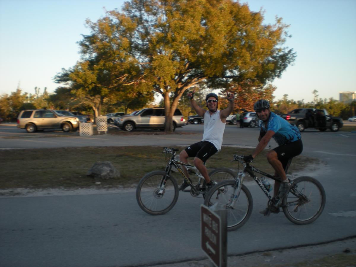 Two men on mountain bikes, smiling and waving, ride along a pathway in a park. In the background, parked cars and trees are visible under a clear sky. Oleta River State Park mountain bike trail.