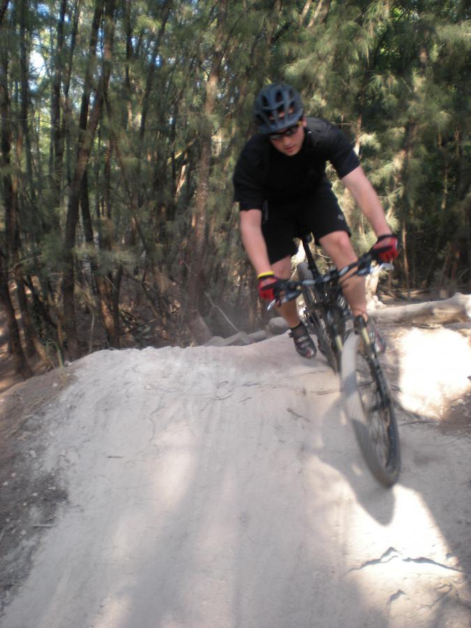 A mountain biker in a black helmet and attire is performing a jump on a dirt trail surrounded by trees. Dust is being kicked up from the ground as the biker navigates a curved section of the path. Oleta River State Park mountain bike trail.