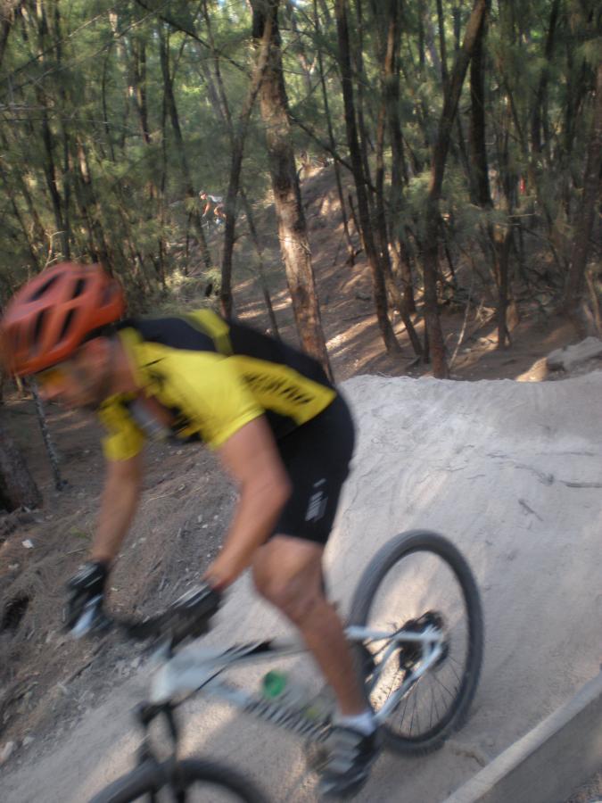A cyclist in a yellow jersey and helmet rides a mountain bike on a dirt trail, surrounded by trees. The image captures the motion and excitement of biking through a forested area. Oleta River State Park mountain bike trail.