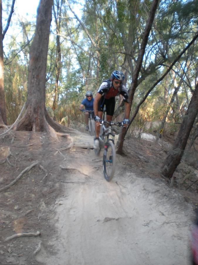 Two mountain bikers navigate a dirt path through a forested area. The first rider, wearing a red and black jersey and a blue helmet, is airborne, while the second rider follows closely behind. Trees and foliage surround the trail, creating a natural setting for outdoor cycling. Oleta River State Park mountain bike trail.