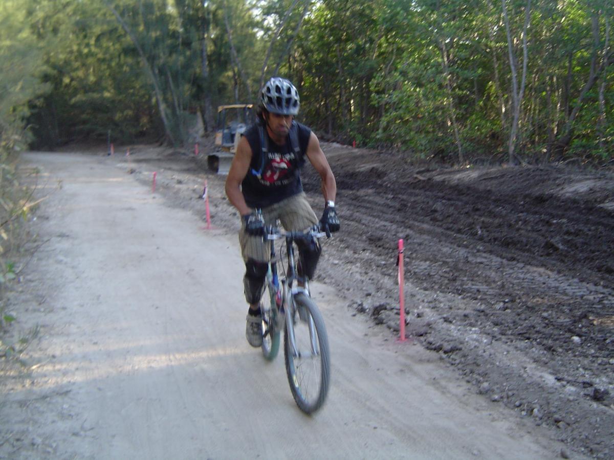 A person riding a mountain bike on a dirt trail surrounded by trees. The cyclist is wearing a helmet, gloves, and knee pads, and is moving along a path marked by pink flags. In the background, a vehicle is parked on the side of the trail. Oleta River State Park mountain bike trail.