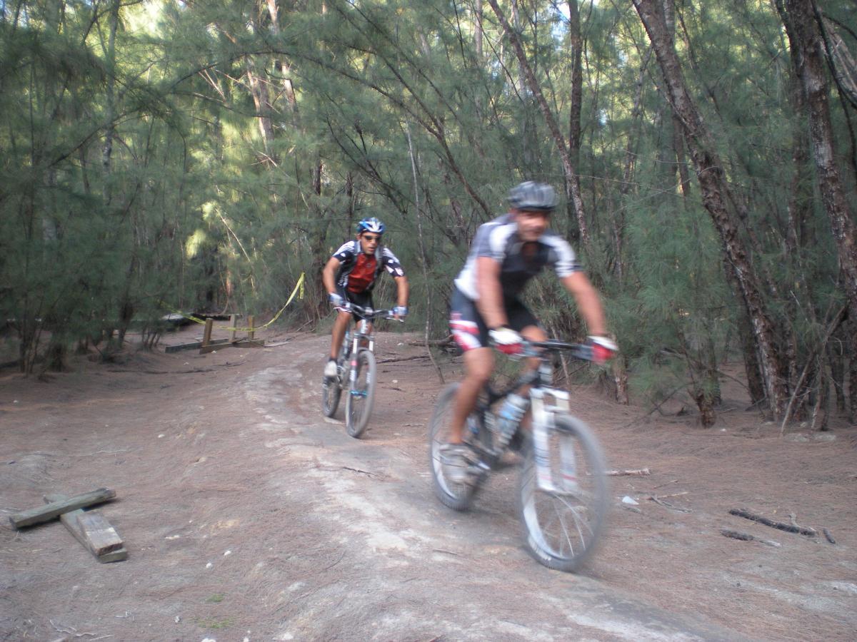 Two mountain bikers riding along a dirt trail in a wooded area, with trees in the background. The riders are in motion, showcasing their biking gear and the rough terrain. The scene captures a sense of adventure and outdoor activity. Oleta River State Park mountain bike trail.