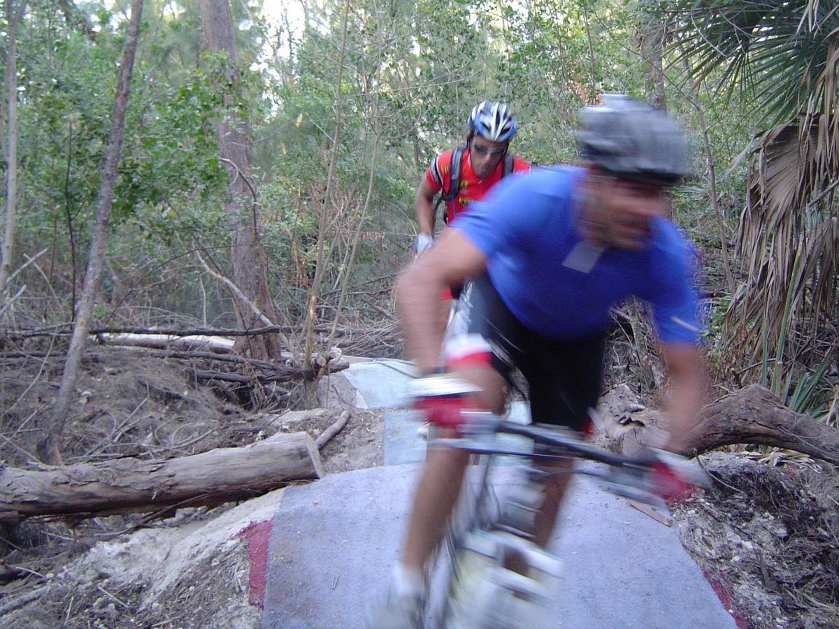 Two mountain bikers navigating a rocky trail through a wooded area. One rider is in sharp focus, moving quickly downhill, while the other rider is slightly blurred in the background. The scene features natural greenery and scattered logs along the path. Oleta River State Park mountain bike trail.