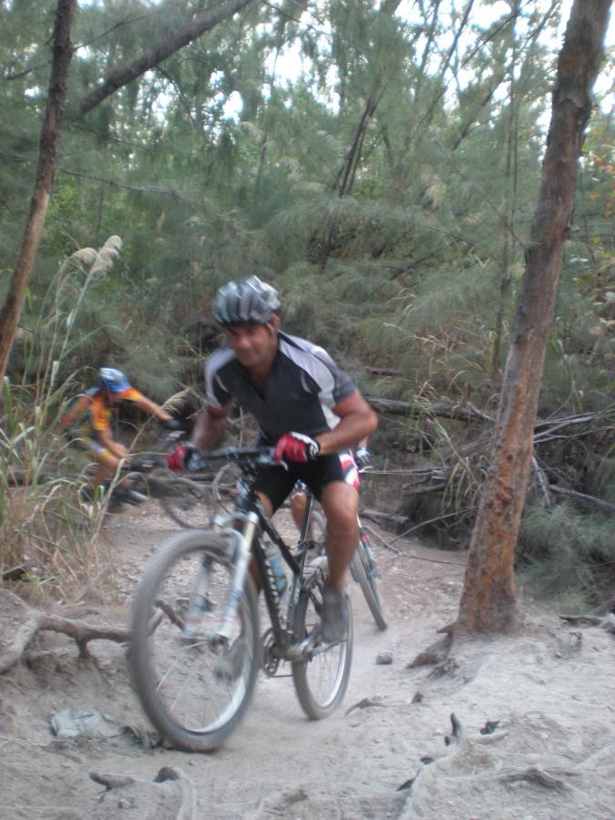 Mountain bikers navigating a rugged trail through a wooded area, with one cyclist in the foreground focused on maintaining balance while riding. The second cyclist is slightly blurred in the background, emphasizing motion. The trail is sandy with roots and greenery surrounding the path. Oleta River State Park mountain bike trail.