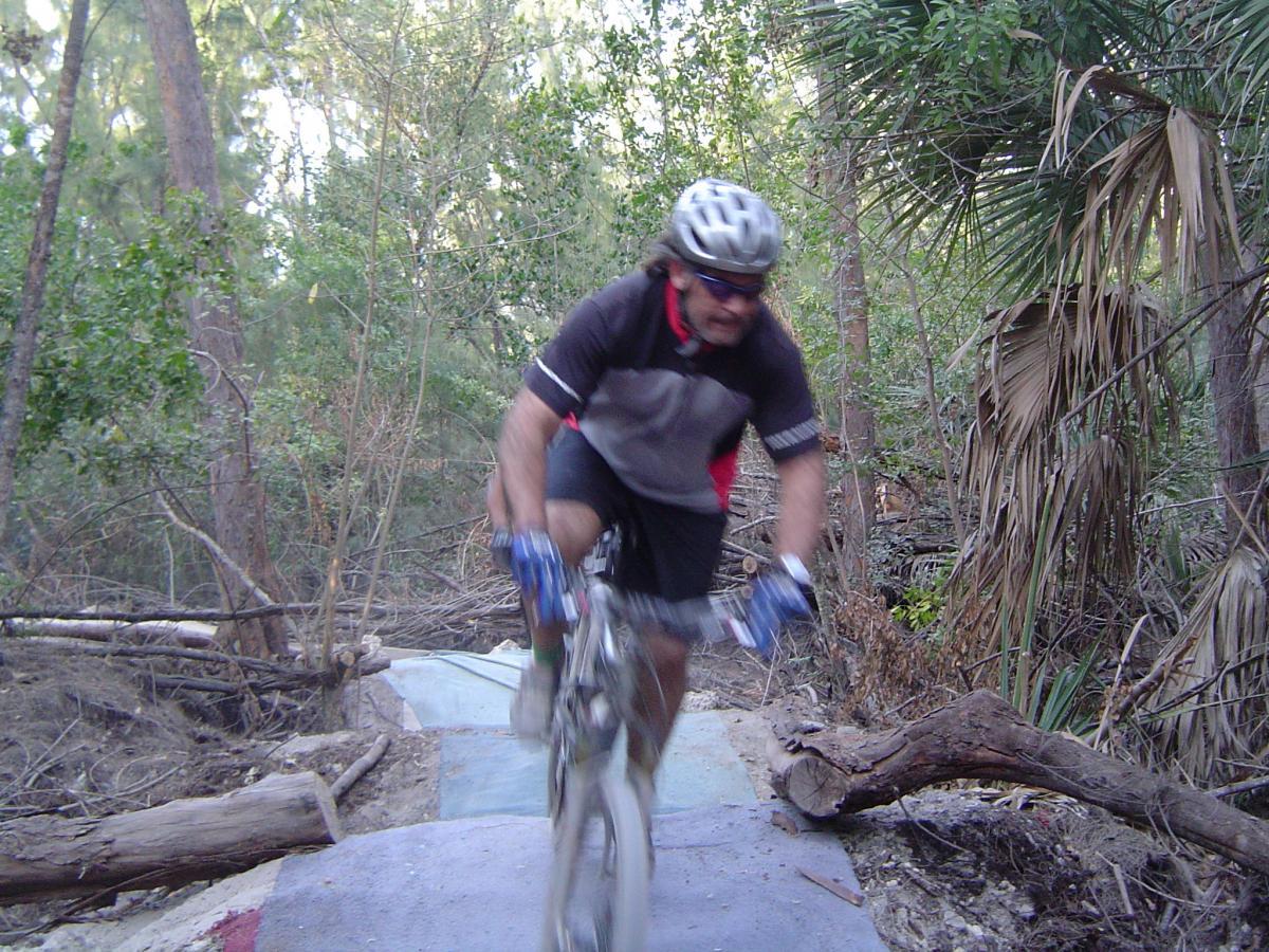 A mountain biker in motion, wearing a helmet and cycling gloves, navigates a rough trail surrounded by trees and vegetation. The bike is airborne as he approaches a jump on the uneven path, showcasing his adventurous spirit and skill. Oleta River State Park mountain bike trail.