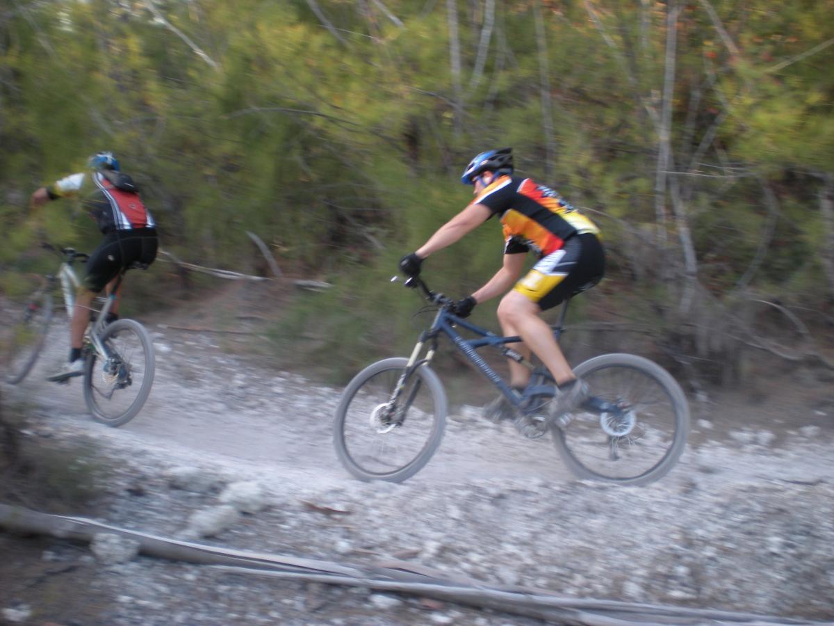Two mountain bikers riding along a dusty trail in a wooded area. One athlete is in a red and black jersey, while the other wears a colorful jersey with yellow and orange. The scene conveys a sense of motion as they navigate the rugged terrain surrounded by trees. Oleta River State Park mountain bike trail.