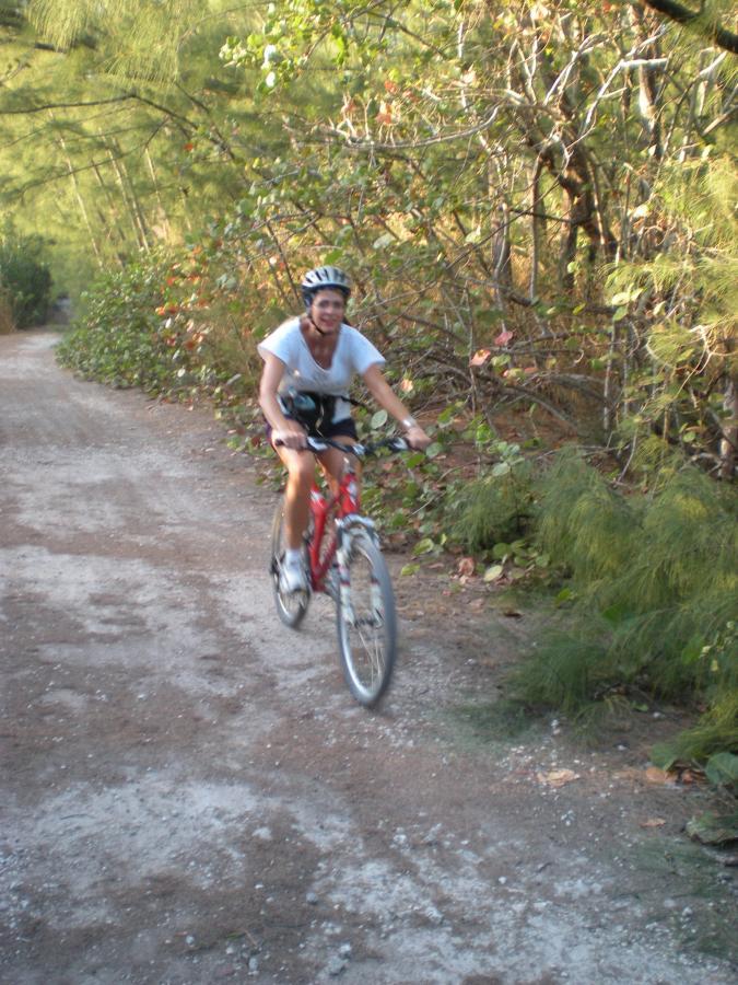 A person riding a red mountain bike along a dirt path surrounded by lush greenery and trees, wearing a helmet and casual clothing. The image captures a sense of outdoor adventure and active lifestyle. Oleta River State Park mountain bike trail.