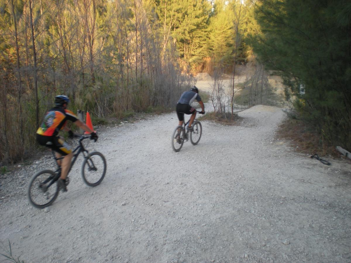 Two mountain bikers ride along a gravel path surrounded by trees, one wearing a brightly colored jersey and the other in a black outfit. There's a traffic cone positioned near the path, indicating a direction or caution for the riders. The setting suggests an outdoor trail suitable for biking. Oleta River State Park mountain bike trail.