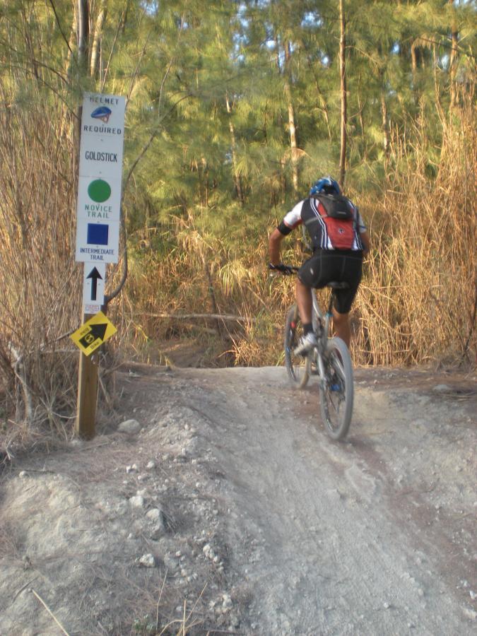 A mountain biker wearing a helmet and backpack rides up a dirt trail surrounded by trees and brush. A trail sign indicating "Helmet Required," "Goldstick," and designations for "Novice Trail" and "Intermediate Trail" is visible in the foreground. Oleta River State Park mountain bike trail.