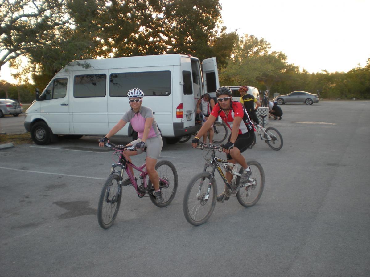 A group of mountain bikers in a parking lot, ready to ride. A woman in a pink cycling outfit smiles as she poses on her pink bike, while a man in a red and black jersey rides beside her. In the background, a van is parked, and other cyclists are preparing for their ride. The scene is set in a natural area during the evening. Oleta River State Park mountain bike trail.