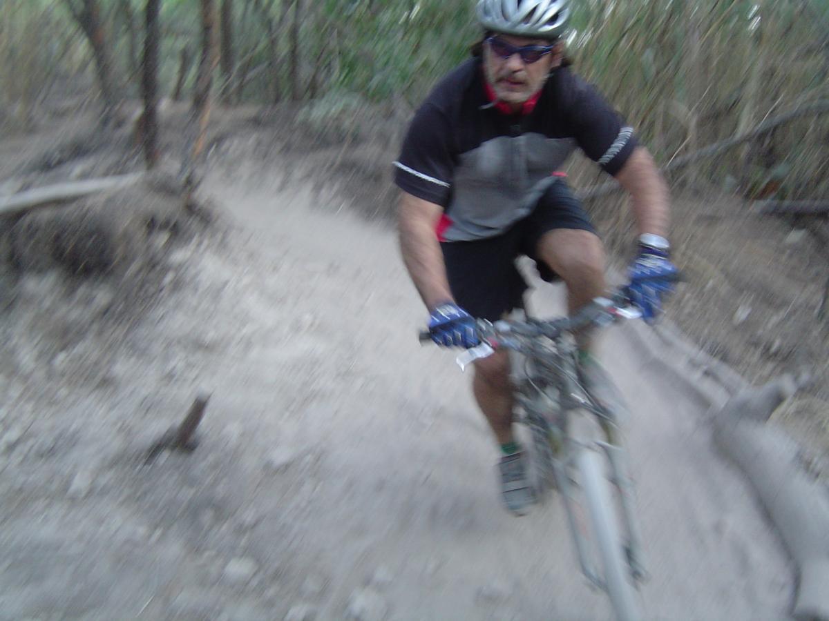 A person riding a mountain bike on a dirt trail surrounded by trees, captured in motion, with a blurred background indicating speed. The cyclist is wearing a helmet, sunglasses, and cycling gloves, appearing focused on navigating the trail. Oleta River State Park mountain bike trail.