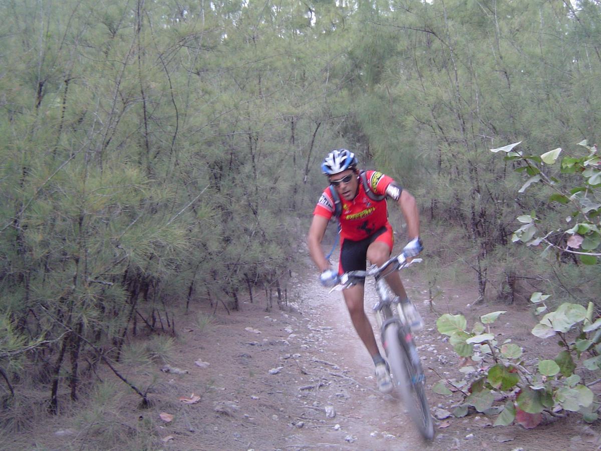 A mountain biker in a red and yellow jersey rides down a gravel trail surrounded by dense green trees. The rider is captured mid-motion, showing focus and determination as he navigates through the natural landscape. Oleta River State Park mountain bike trail.