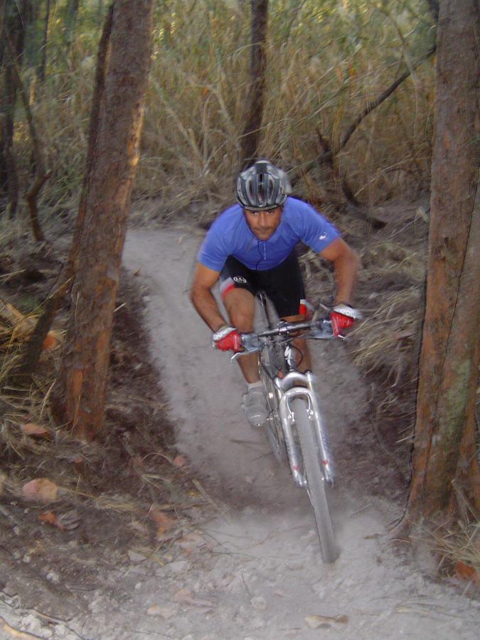 A mountain biker navigating a narrow dirt trail surrounded by trees, wearing a blue shirt and a helmet, with dust rising from the ground as he rides. Oleta River State Park mountain bike trail.