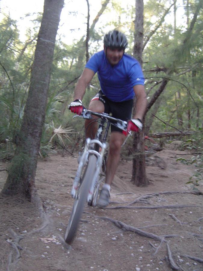 A person riding a mountain bike on a dirt trail surrounded by trees, with a motion blur suggesting speed. The rider is wearing a blue shirt and a helmet, focusing on navigating the uneven terrain. Oleta River State Park mountain bike trail.