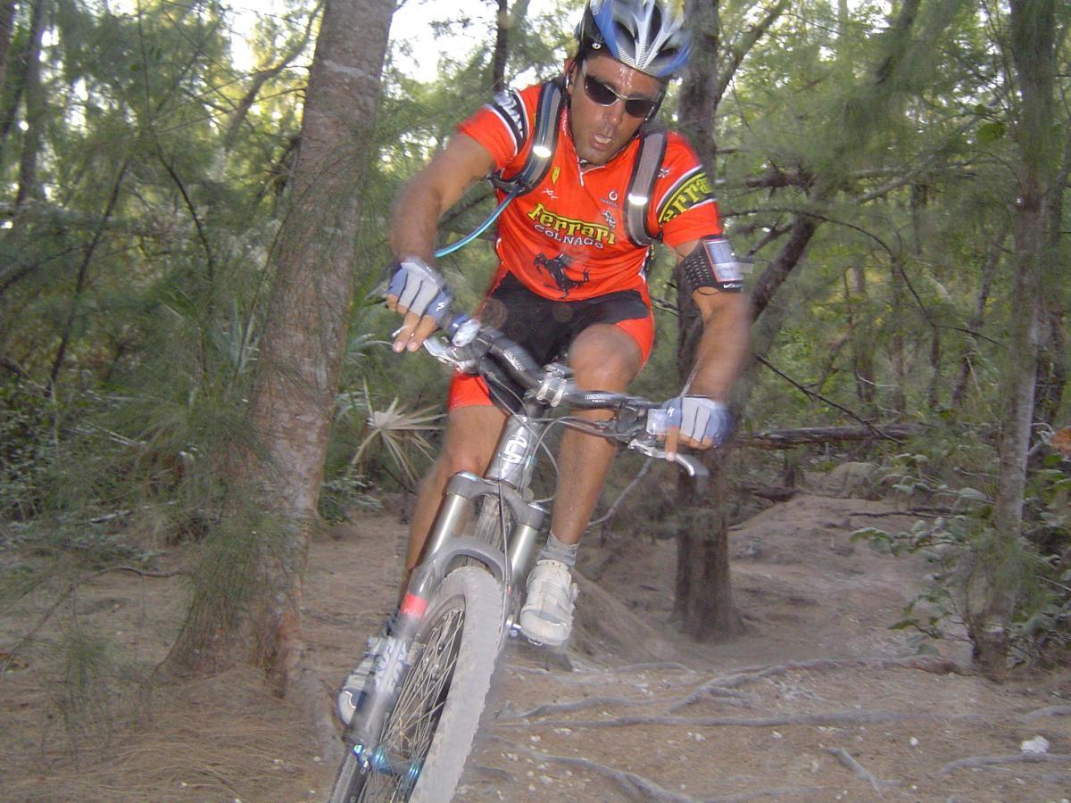 A mountain biker in an orange jersey and helmet rides swiftly through a forest trail, navigating around trees and uneven terrain. The rider appears focused and determined, capturing the excitement of outdoor cycling. Oleta River State Park mountain bike trail.