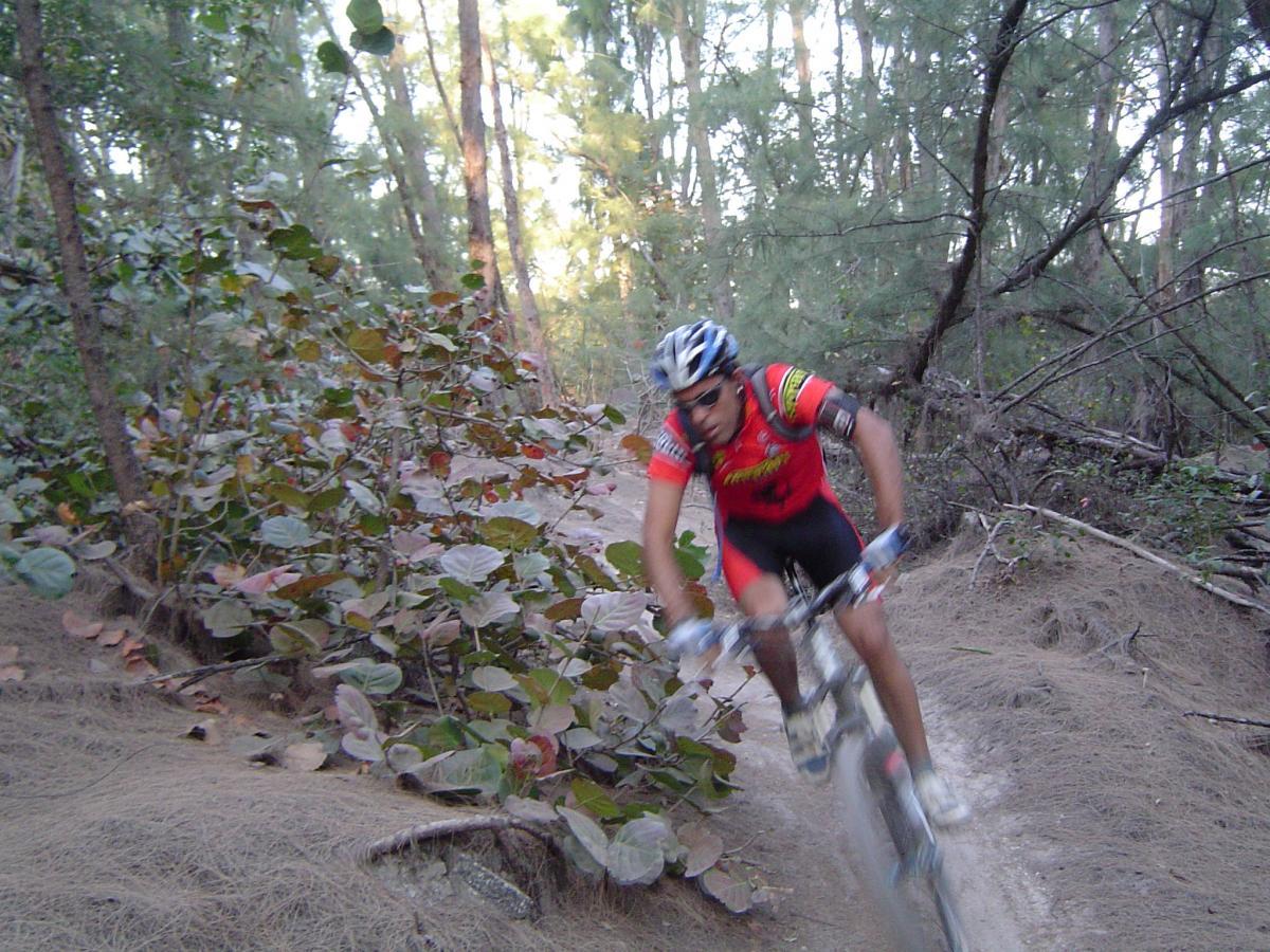 A mountain biker navigating a dirt trail through a wooded area, surrounded by trees and bushes. The cyclist is in motion, wearing a red and black jersey and helmet, with dirt and vegetation visible along the path. Oleta River State Park mountain bike trail.