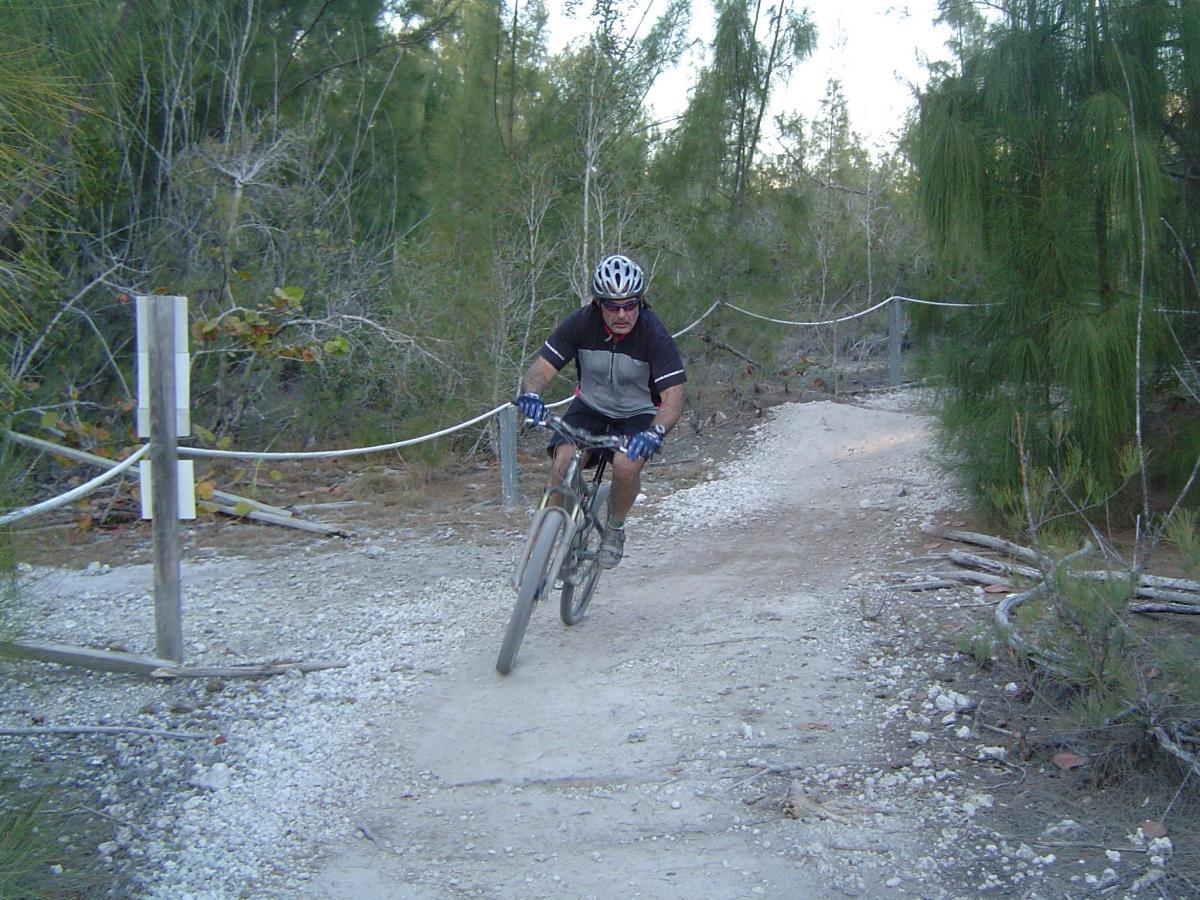A mountain biker navigating a narrow, rocky trail surrounded by trees. The rider is wearing a helmet and gloves, leaning forward as they maneuver the bike along the path. A rope boundary is visible on the left side of the image. Oleta River State Park mountain bike trail.