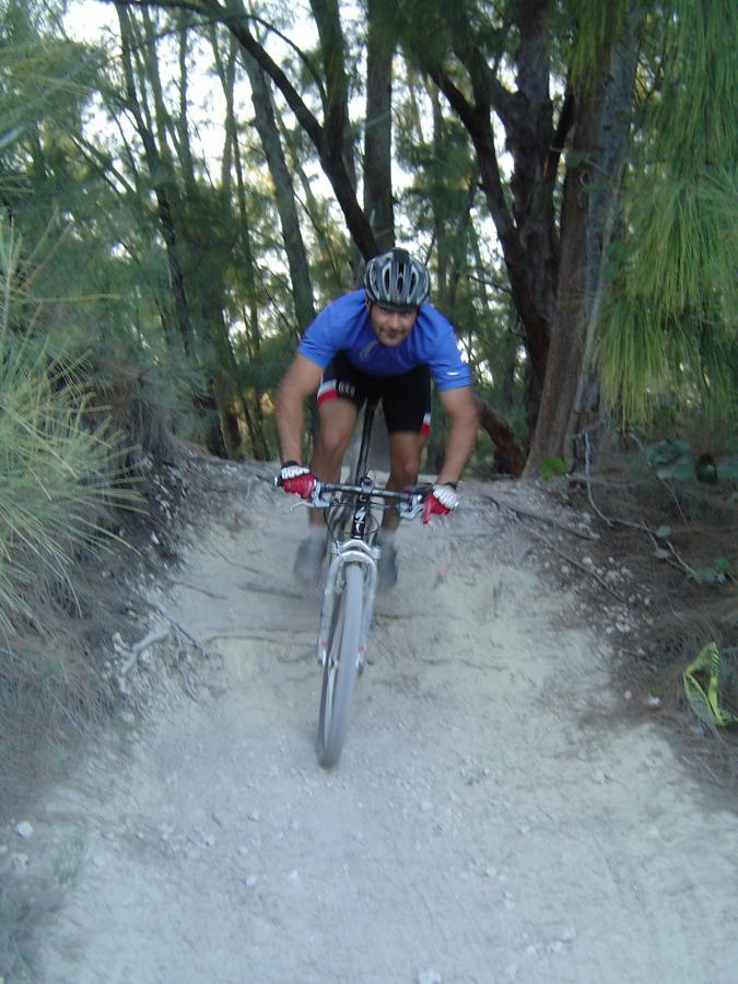 A mountain biker in a blue shirt and black shorts navigates a sandy trail surrounded by trees, showcasing agility and focus while riding downhill. Oleta River State Park mountain bike trail.