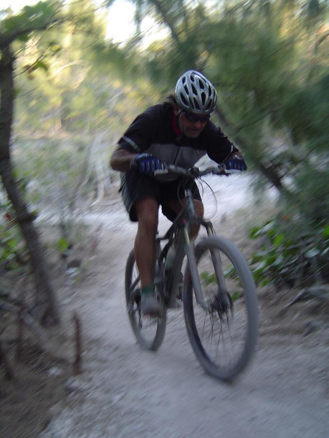 A person riding a mountain bike on a dirt trail surrounded by greenery, captured in motion with a slight motion blur to convey speed. Oleta River State Park mountain bike trail.