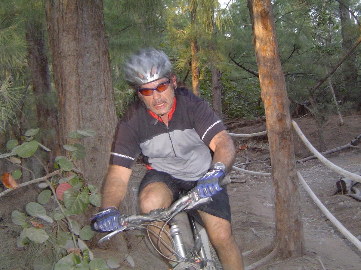 A mountain biker wearing a helmet and sunglasses navigates a forest trail, surrounded by trees and vegetation. The biker is focused and exerting effort while riding on a dirt path. Oleta River State Park mountain bike trail.