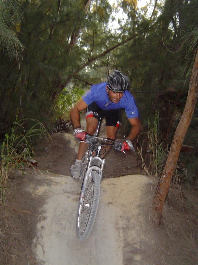 A mountain biker navigating a dirt trail surrounded by trees, leaning forward as he maneuvers over a small jump. He is wearing a blue shirt, black shorts, and a helmet, with his bike partially in the air. Oleta River State Park mountain bike trail.