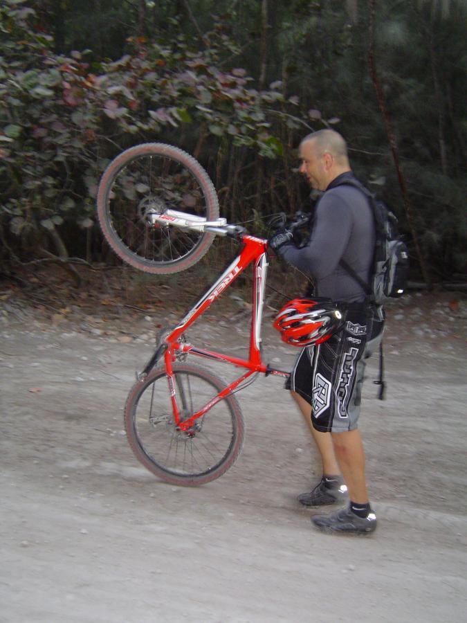 A person in a black shirt and cycling shorts is walking on a dirt path, holding a red mountain bike by the front tire. They are wearing a helmet and a backpack, with trees and foliage in the background. Oleta River State Park mountain bike trail.