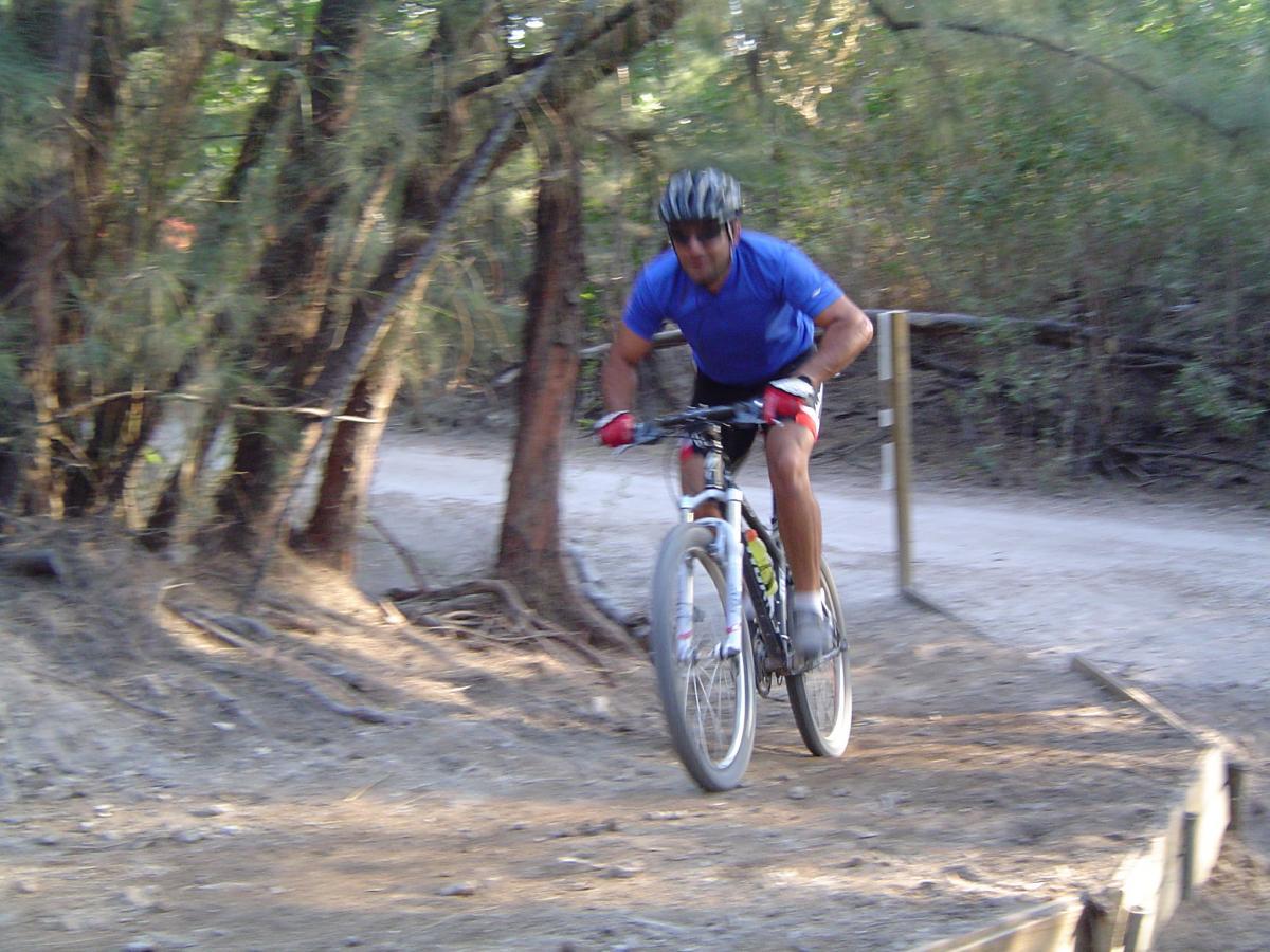 A cyclist in a blue shirt rides a mountain bike over a dirt path, surrounded by trees. The rider appears to be in motion, leaning forward, and navigating a rustic trail. Oleta River State Park mountain bike trail.