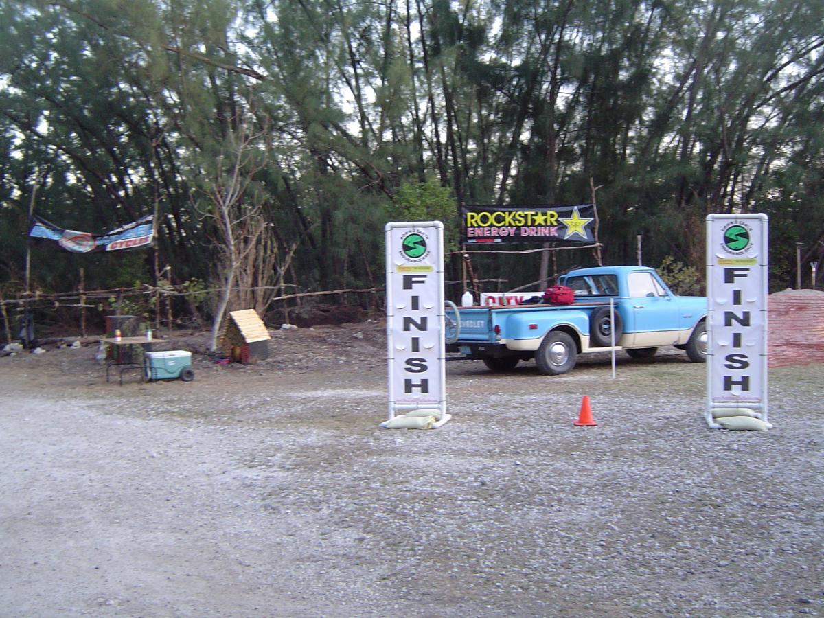 A finish line setup at an outdoor event, featuring two tall signs labeled "FINISH" in bold letters. In the background, a blue vintage truck is parked near a promotional banner for Rockstar Energy Drink. Nearby, there is a small table and a cooler, alongside a patch of trees. The ground is gravel, and an orange cone is placed at the finish line. Oleta River State Park mountain bike trail.