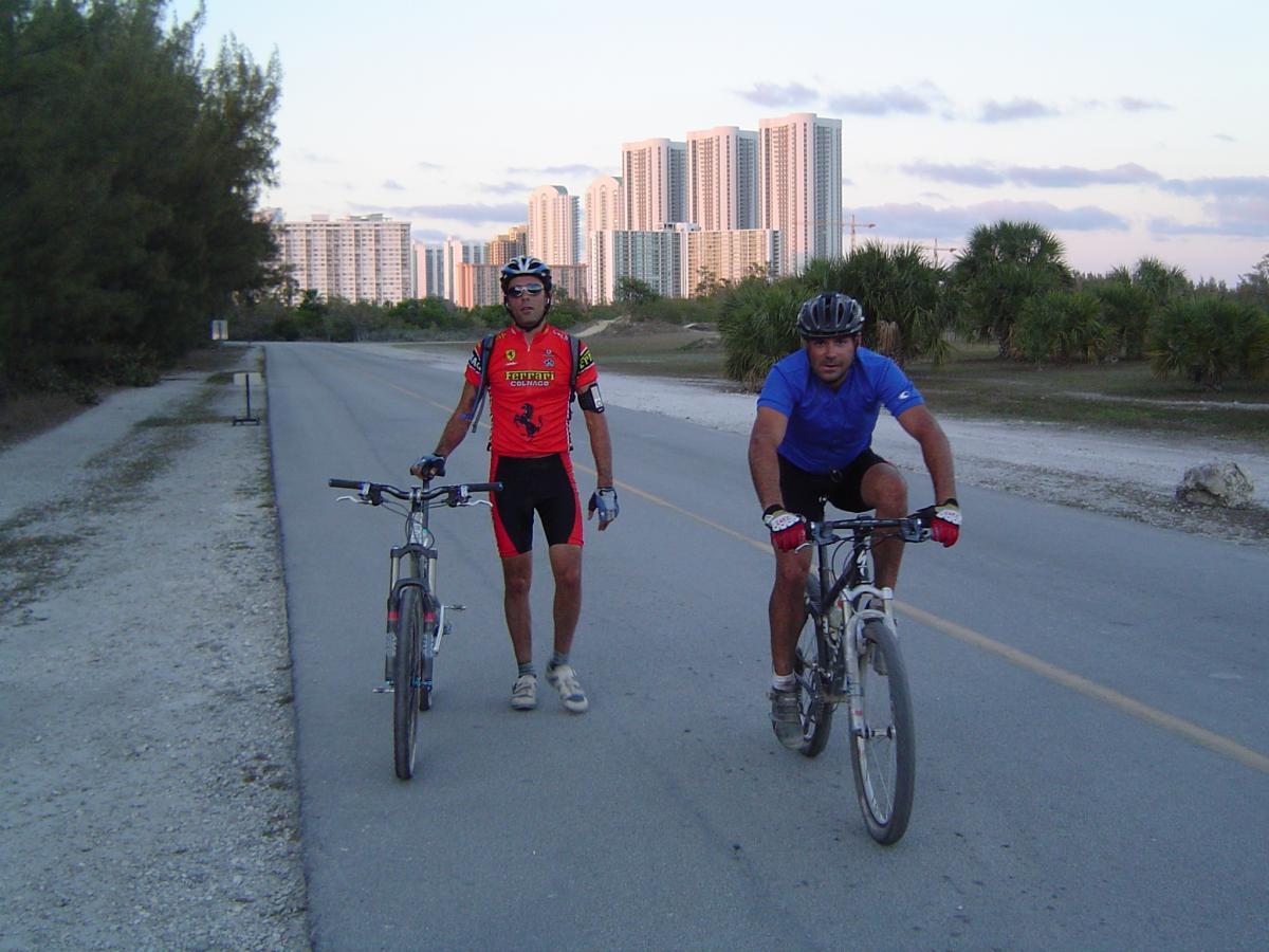 Two men are on a paved road, one standing beside his bicycle and the other riding his bike. The first man wears a red cycling jersey with black shorts and sunglasses, while the second man is in a blue shirt and black shorts with gloves. In the background, there are tall buildings and palm trees under a cloudy sky. Oleta River State Park mountain bike trail.