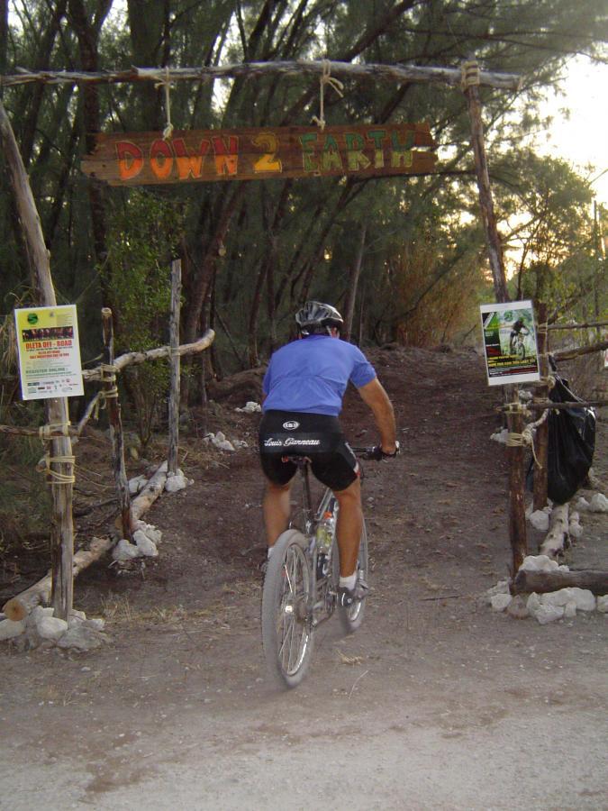 A cyclist riding a mountain bike toward a wooden archway with a sign that reads "DOWN 2 EARTH," surrounded by trees and a dirt path, with various flyers posted nearby. Oleta River State Park mountain bike trail.