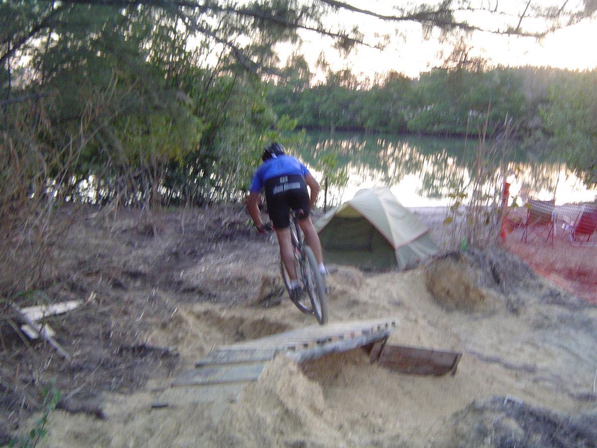 A mountain biker wearing a blue jersey is jumping off a small ramp made of wooden planks, with a sandy area and a river visible in the background. A camping tent is set up nearby, surrounded by trees. The scene is captured during sunset, casting a warm glow over the landscape. Oleta River State Park mountain bike trail.
