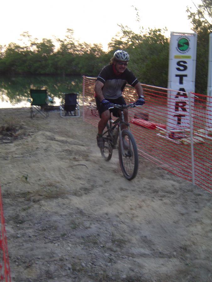 A mountain biker in a helmet rides along a dirt path near a body of water, approaching the start line marked by a visible banner reading "START." Surrounding the area are chairs and safety fencing, with trees in the background and a soft evening light illuminating the scene. Oleta River State Park mountain bike trail.