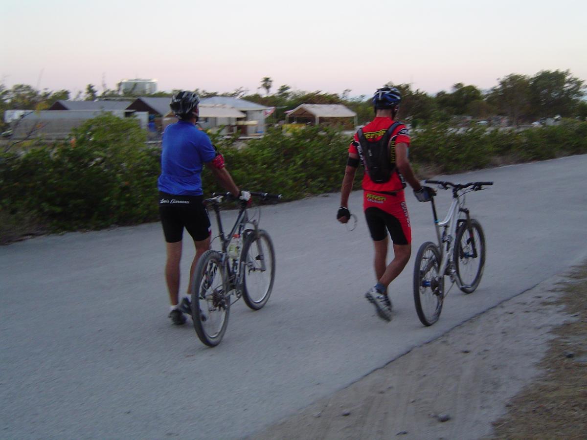 Two cyclists walking their mountain bikes along a dirt path, surrounded by greenery and buildings in the background. One cyclist is wearing a blue jersey and black shorts, while the other is dressed in a red jersey with a black backpack. The scene is set in the early evening, with soft light illuminating the surroundings. Oleta River State Park mountain bike trail.