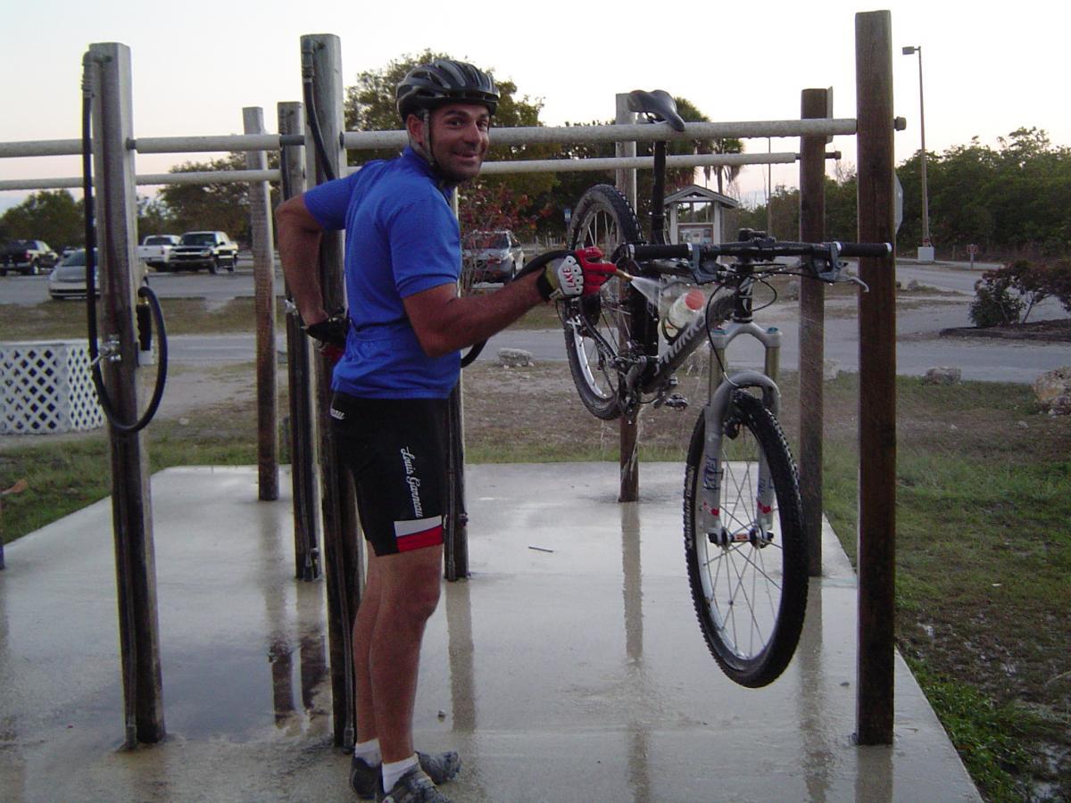 A cyclist in a blue shirt and black shorts smiles while preparing to wash a mountain bike at a bike cleaning station. The bike is suspended from a horizontal bar, and the area is outdoors with a parking lot visible in the background. The sun is setting, casting a warm light over the scene. Oleta River State Park mountain bike trail.