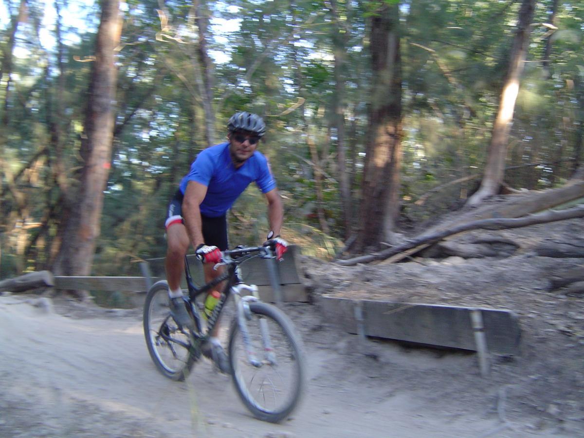 A person riding a mountain bike on a sandy trail surrounded by trees. The rider is wearing a blue shirt, black shorts, and a helmet. The image captures the motion and excitement of mountain biking in a natural setting. Oleta River State Park mountain bike trail.