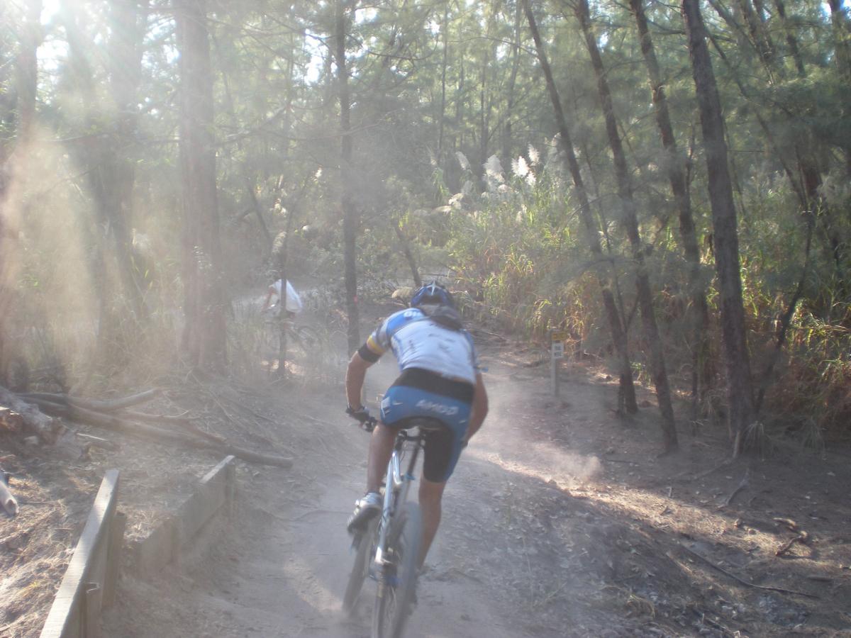 A mountain biker riding on a dusty trail through a forested area, with sunlight filtering through the trees, creating a misty atmosphere. Another cyclist is seen in the background. Oleta River State Park mountain bike trail.