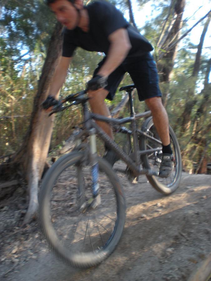 A cyclist riding a mountain bike over a dirt path in a wooded area, captured in motion with a blurred effect. The cyclist is wearing a black shirt and shorts, demonstrating an active stance as they navigate the trail. Trees and foliage are visible in the background, indicating a natural outdoor setting. Oleta River State Park mountain bike trail.