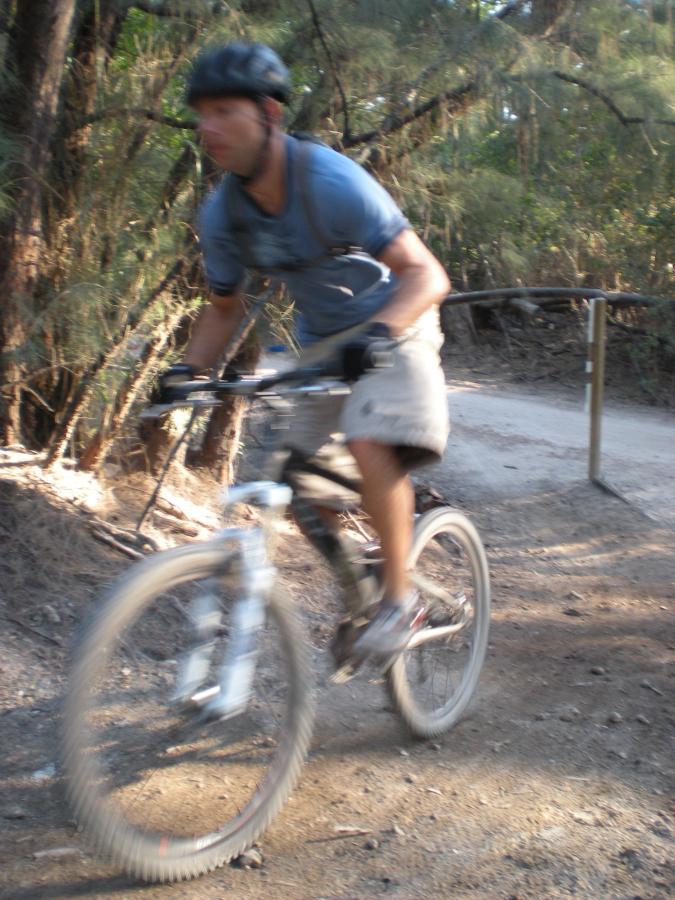 A blurred image of a person riding a mountain bike along a dirt trail surrounded by trees, capturing the motion and speed of the cyclist. Oleta River State Park mountain bike trail.